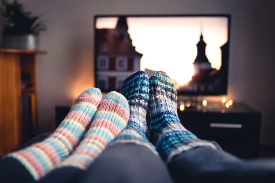 Two people in knitted socks with their feet up in front of a television