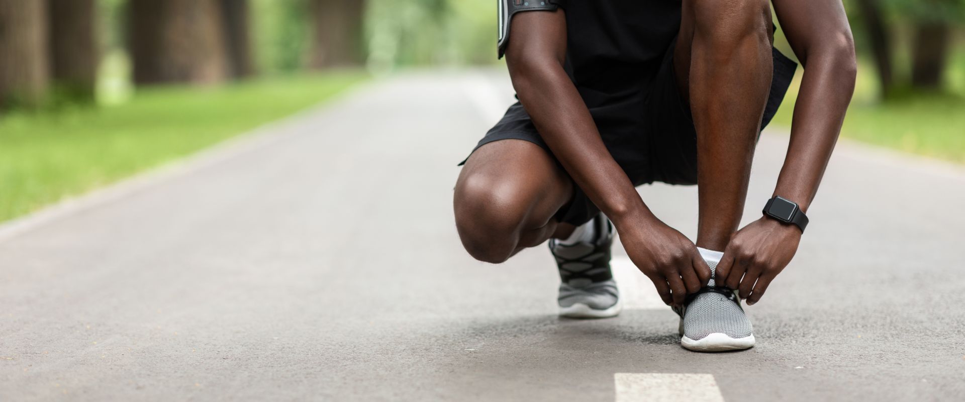 Close-up shot of a male runner crouched down on a paved path in an outdoor park, tying the laces of his grey running shoe before a run.