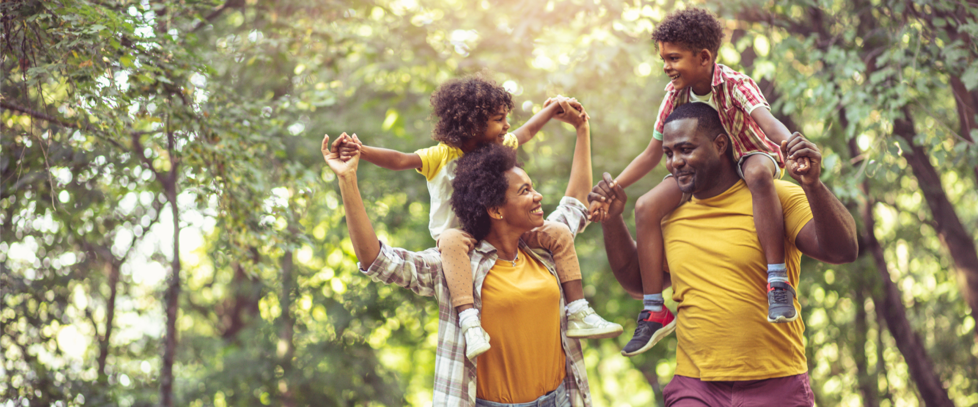 A happy family of four walking through a park or wooded area during daytime. The mother and father are carrying their young daughter and son on their shoulders while holding hands.