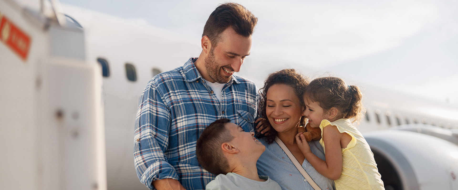 A happy family of four standing together and smiling outside on an airport tarmac next to a white airplane before boarding.