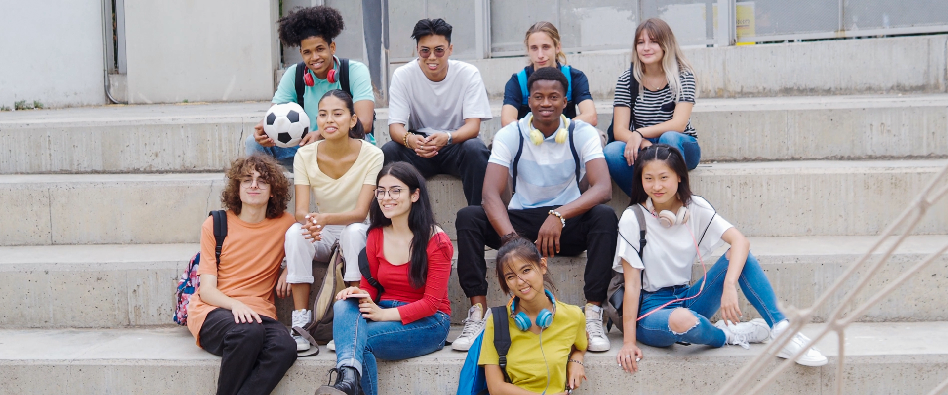 Group portrait of a diverse group of ten young students sitting together outdoors on wide concrete steps outside a university or school building. Several students are carrying backpacks, wearing headphones around their necks, and one student holds a soccer ball.