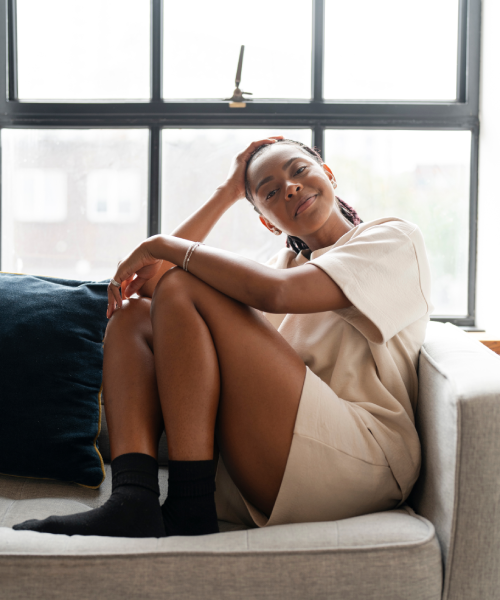 Portrait of a young woman resting comfortably on a grey couch, sitting with her knees pulled up to her chest and one hand resting gently behind her head, smiling softly at the camera.