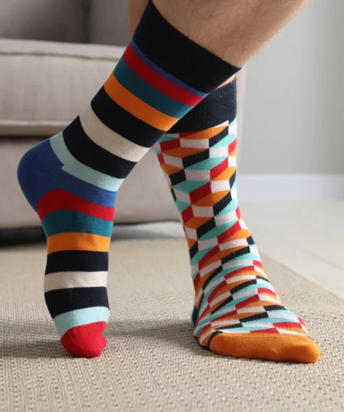 Close-up shot focusing on a person's feet and lower legs wearing a pair of mismatched, brightly colored crew socks indoors. One sock features a horizontal striped pattern in dark blue, light blue, orange, red, black, and white; the other sock features a geometric 3D-cube pattern in similar vibrant colors with an orange toe.